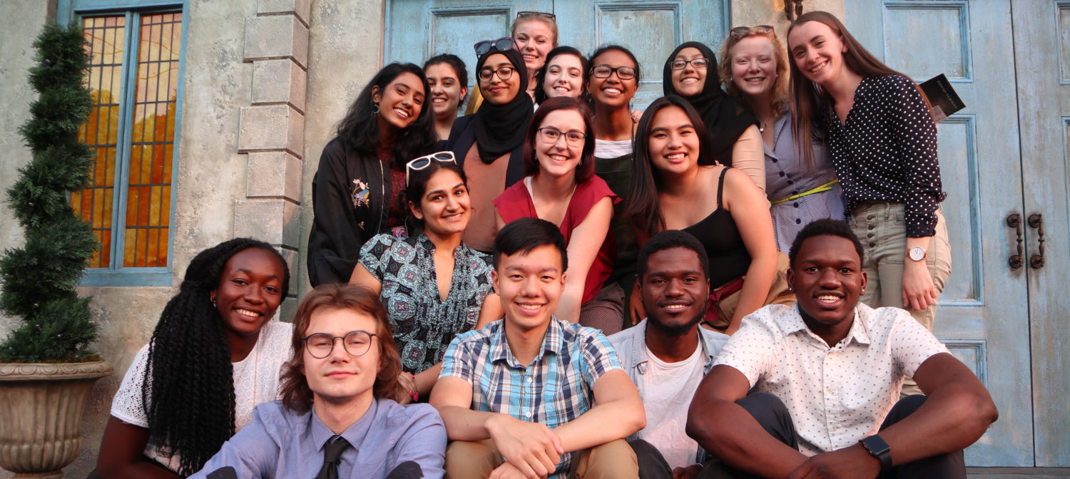 A group shot of Watson fellows. They sit and stand on steps, smiling at the camera, blue doors and green foliage behind them