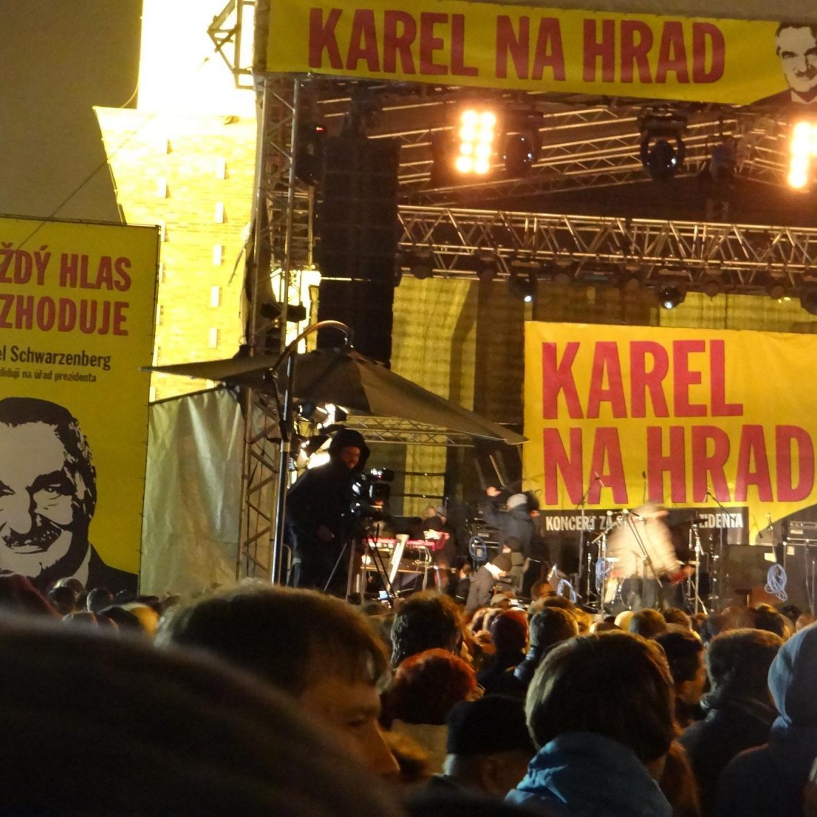 outdoor concert for a Czech political campaign, man on stage with a microphone in hand and mixtables beside him, posters around in yellow