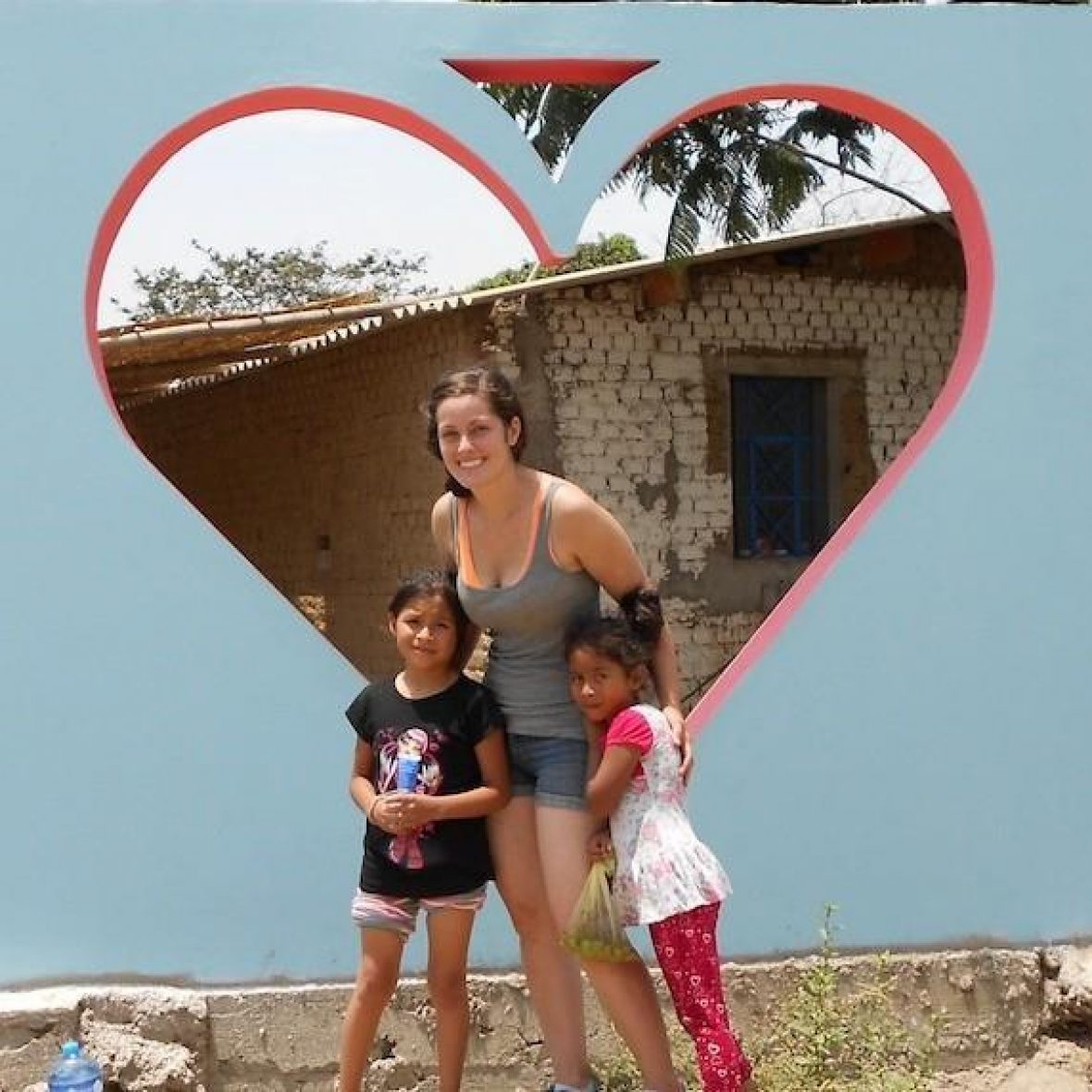 MaryBeth Apriceno wearing a tank top and denim shorts smiles at the camera, hugging a child with each arm, with graffiti in the shape of a heart on the wall behind her