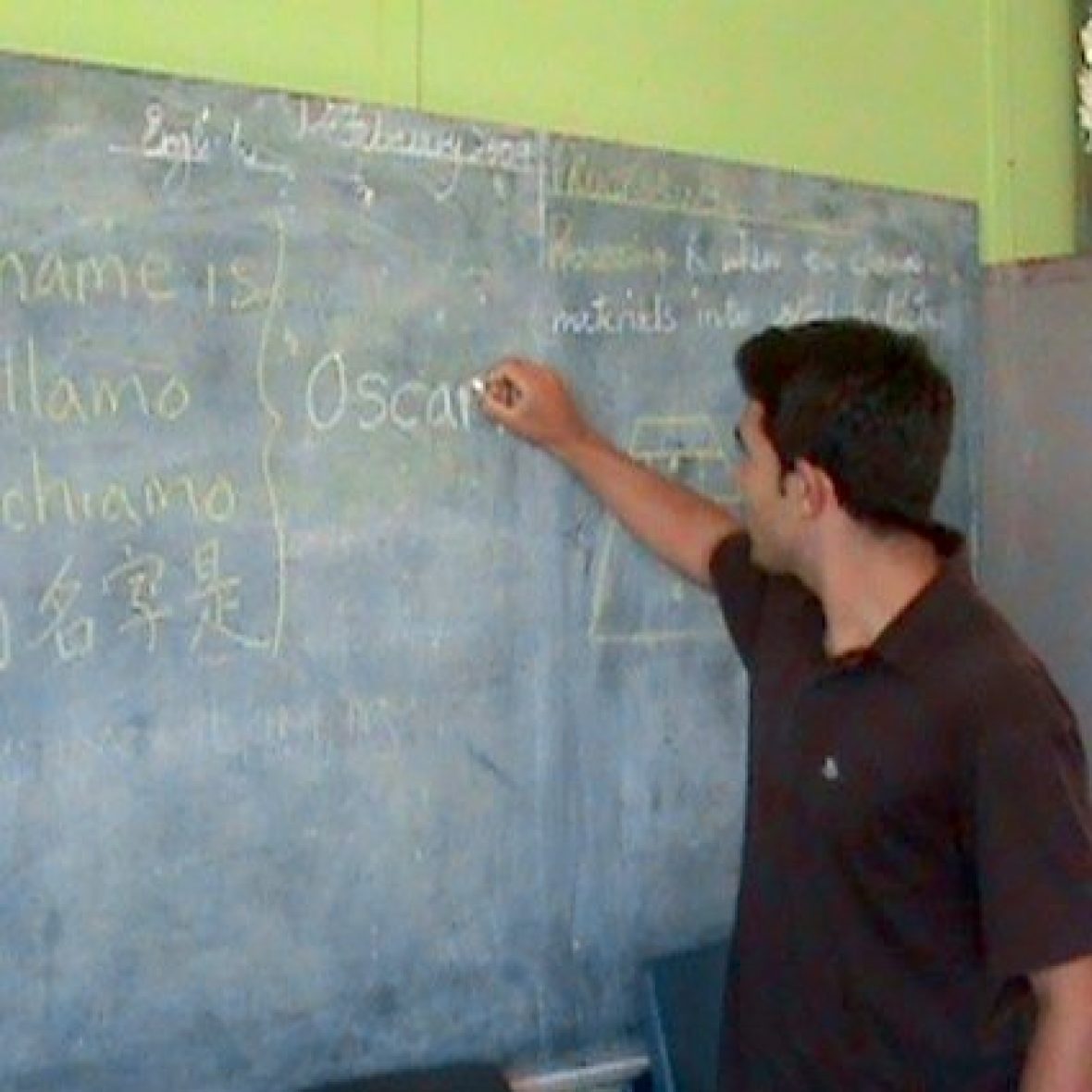 Oscar Baez with his back to the camera and wearing a black shirt, writing on a blackboard in white chalk with words in Spanish on the board