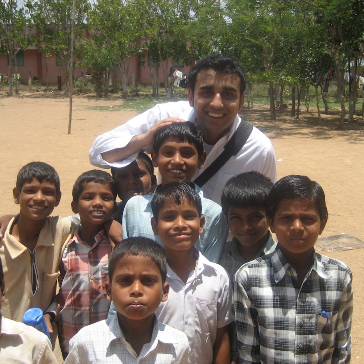 Reggie Thomas, wearing a white button-down with a camera strap across his chest, smiles at the camera from behind a crowd of smiling children, beige earth and green trees in the background