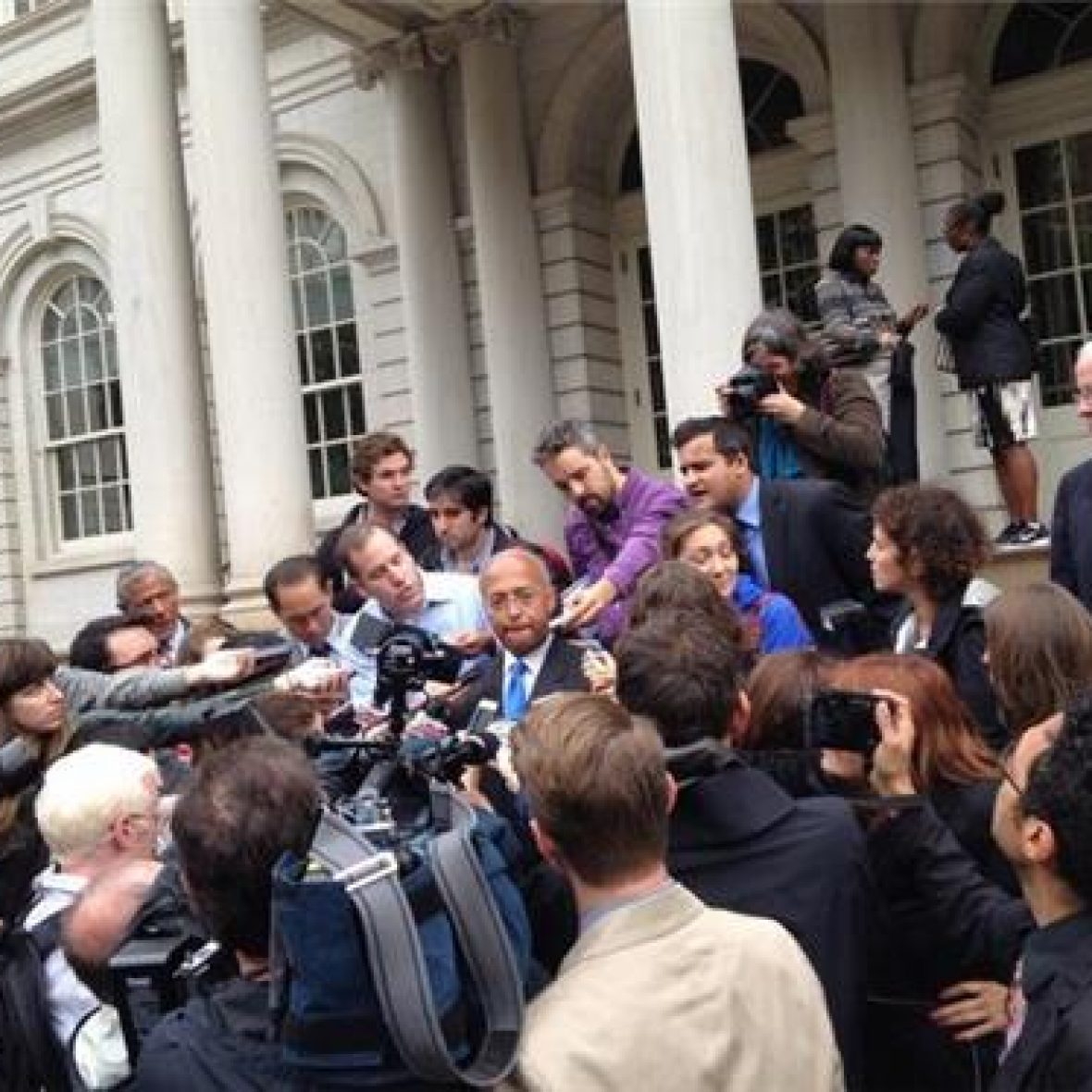 politician stands at the center of a crowd at a press conference, speaking into a microphone, journalists and observers clustered around, columns of building in the background